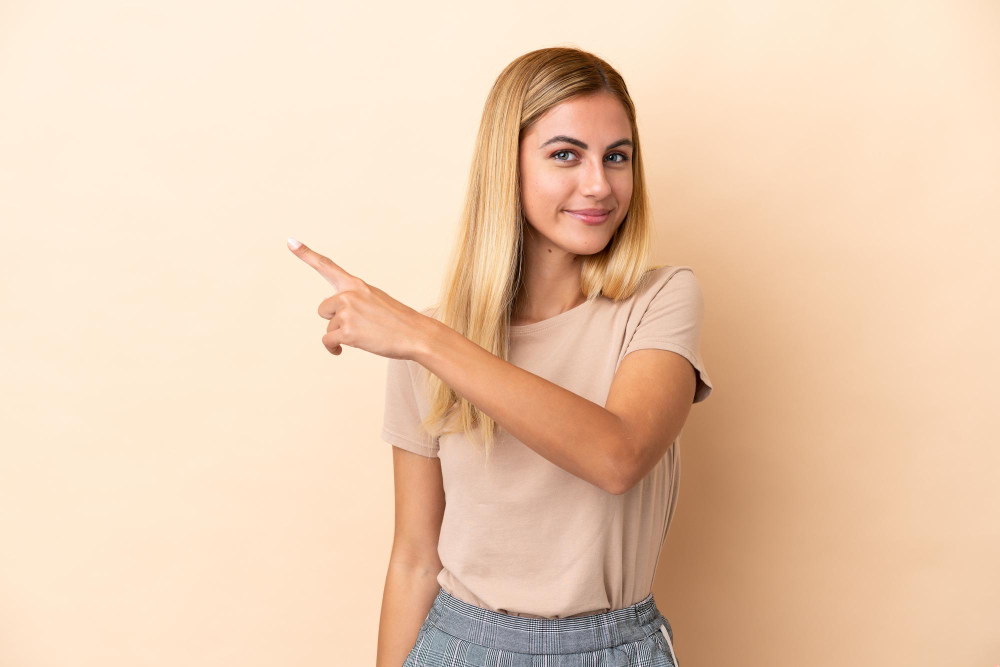 Uma mulher com longos cabelos louros está a sorrir e a apontar para a direita, num fundo bege. Está vestida com uma camisa bege e uma saia axadrezada cinzenta.